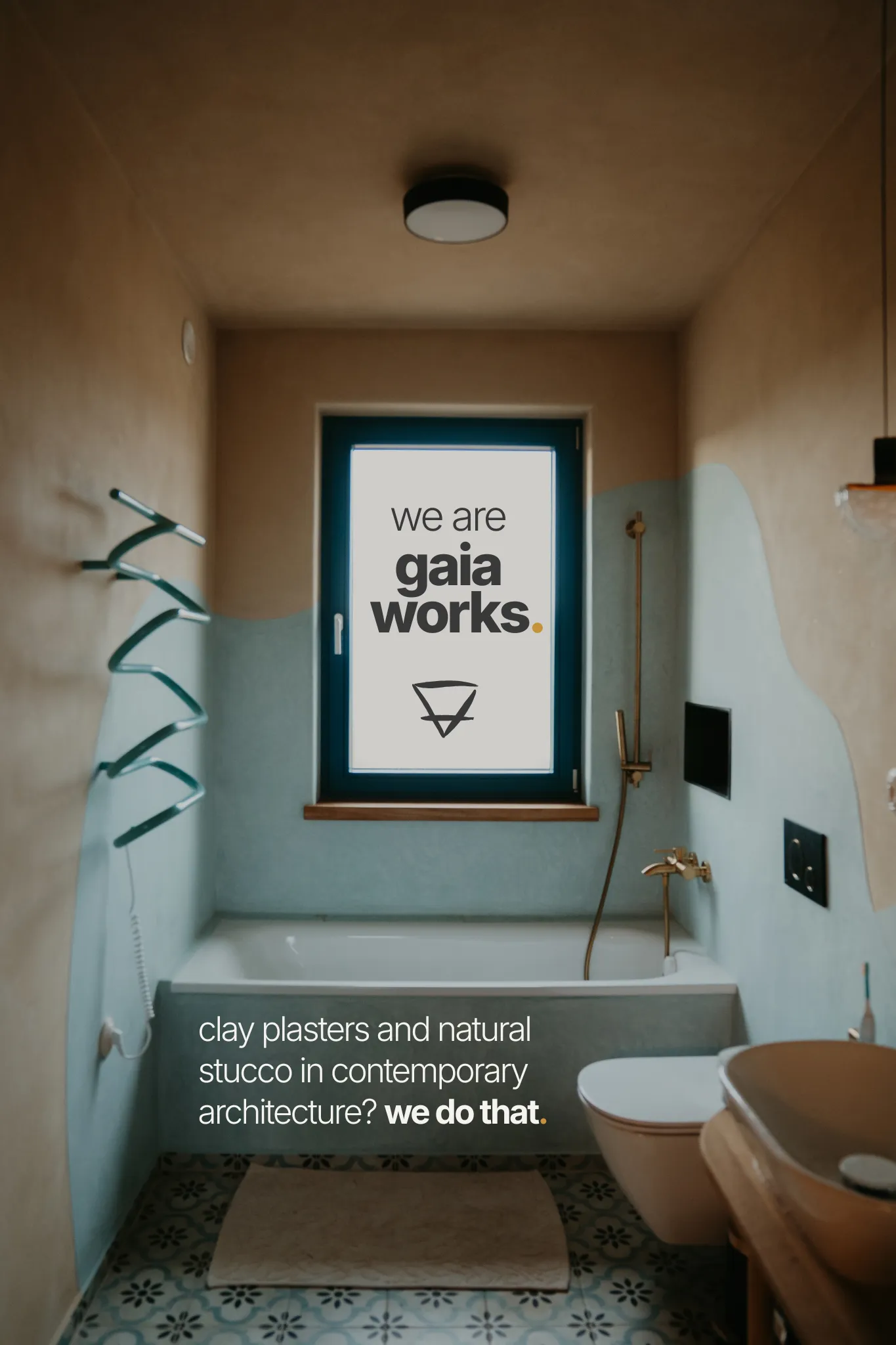 A tadelakt bathroom with a bright window, the text 'We are GAIA WORKS' and the earth symbol in the window, and the text 'Clay plasters and natural stucco in contemporary architecture? We do that.' on the tub.